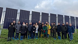 Gruppe von Menschen mit blauen Überschuhen steht vor mehreren großen Solarpanelen auf einer Wiese unter bewölktem Himmel.