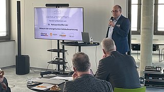 Mann im Anzug hält Mikrofon und spricht vor Bildschirm mit Text zu einem Netzwerktreffen, mehrere Personen sitzen an Tischen und hören zu.