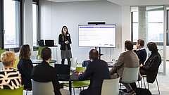 Sieben Personen sitzen auf Stühlen und schauen nach vorne, wo eine Frau einen Vortrag vor einem Bildschirm hält