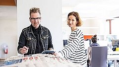 Zwei Personen stehen in einem Büro mit Zeitungen.