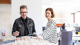 Zwei Personen stehen in einem Büro mit Zeitungen.