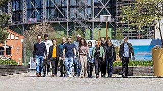 Gruppe von elf Personen unterschiedlichen Alters und Geschlechts, die auf einem gepflasterten Weg in einem urbanen Bereich mit Baustelle im Hintergrund gehen