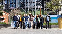 Gruppe von elf Personen unterschiedlichen Alters und Geschlechts, die auf einem gepflasterten Weg in einem urbanen Bereich mit Baustelle im Hintergrund gehen