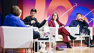 Vier Personen sitzen auf weißen Sesseln auf einer Bühne, eine Person trägt eine schwarze Kappe und gestikuliert mit der Hand, im Hintergrund bunte Wand mit Pfeil- und Zahlenmotiven.