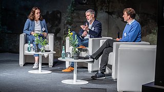 Drei Personen auf der Bühne, die miteinander sprechen.