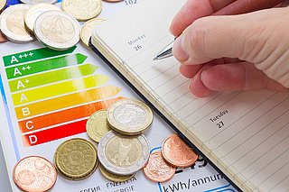 Hand schreibt in einen Kalender, daneben Euro-Münzen und ein Energielabel mit Effizienzklassen von A+++ bis D.