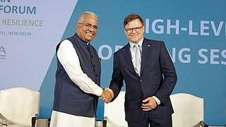 Bundesumweltminister Carsten Schneider und sein indischer Amtskollege Bhupender Yadav geben sich auf der Bühne des 4. Deutsch-Indischen Umweltforums die Hand. Im Hintergrund ist eine Leinwand sichtbar.