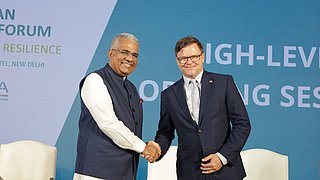 Bundesumweltminister Carsten Schneider und sein indischer Amtskollege Bhupender Yadav geben sich auf der Bühne des 4. Deutsch-Indischen Umweltforums die Hand. Im Hintergrund ist eine Leinwand sichtbar.