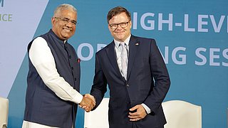 Bundesumweltminister Carsten Schneider und sein indischer Amtskollege Bhupender Yadav geben sich auf der Bühne des 4. Deutsch-Indischen Umweltforums die Hand. Im Hintergrund ist eine Leinwand sichtbar.