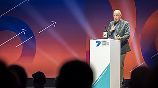 Mann in grauem Sakko steht am Rednerpult mit Mikrofon und Weinflasche, im Hintergrund bunte Kreise mit weißen Pfeilen.