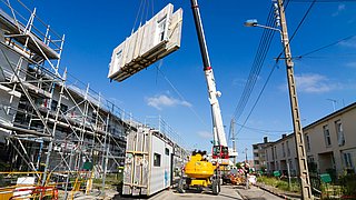 Ein Kran hebt ein großes Bauelement auf einer Baustelle an. Im Hintergrund sind Gerüste und mehrere Gebäude sichtbar. Baumaschinen stehen auf der Straße.