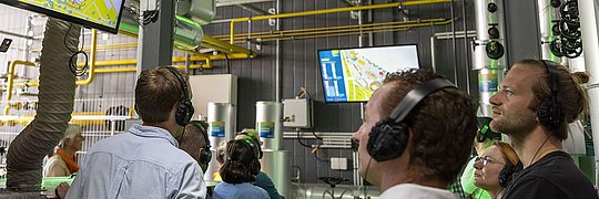  Personen sehen sich ein Video auf einem großen Bildschirm in einer Fabrik an.
