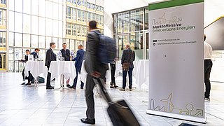 Im Foyer des Hauses der Deutschen Wirtschaft läuft ein Mann in Anzug  vor einem großem Banner der Marktoffensive. Im Hintergrund sind Stehtische mit weiteren Besucher:innen zu sehen. 