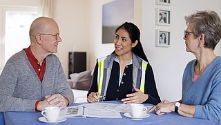 Drei Personen sitzen an einem Tisch mit Bauplänen und Kaffeetassen und führen ein Gespräch