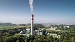 Industrieanlage mit einem hohen Schornstein, aus dem Rauch aufsteigt, umgeben von grüner Landschaft und Feldern. Im Hintergrund sind Hügel und eine Stadt sichtbar. Der Schornstein trägt die Aufschrift "KVA".