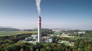 Industrieanlage mit einem hohen Schornstein, aus dem Rauch aufsteigt, umgeben von grüner Landschaft und Feldern. Im Hintergrund sind Hügel und eine Stadt sichtbar. Der Schornstein trägt die Aufschrift "KVA".