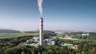 Industrieanlage mit einem hohen Schornstein, aus dem Rauch aufsteigt, umgeben von grüner Landschaft und Feldern. Im Hintergrund sind Hügel und eine Stadt sichtbar. Der Schornstein trägt die Aufschrift "KVA".