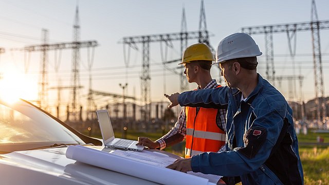 Zwei Bauarbeiter in Schutzhelmen stehen an einem Auto. Einer zeigt auf einen Laptop, während der andere Pläne auf einem Tisch ausbreitet. Im Hintergrund sind Strommasten sichtbar.