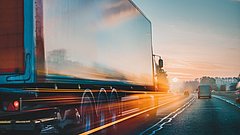 Ein Lkw fährt über eine leere Autobahnspur, die von der Sonne beleuchtet wird. Die Landschaft ist herbstlich.  