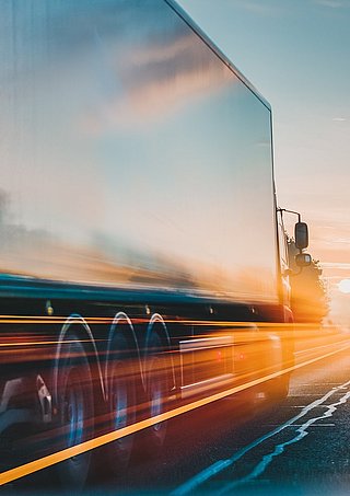Ein Lkw fährt über eine leere Autobahnspur, die von der Sonne beleuchtet wird. Die Landschaft ist herbstlich.  