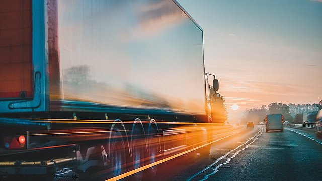 Ein Lkw fährt über eine leere Autobahnspur, die von der Sonne beleuchtet wird. Die Landschaft ist herbstlich.  