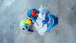 Drei Bauarbeiter mit Schutzhelmen und Warnwesten betrachten einen Bauplan auf einem Betonboden