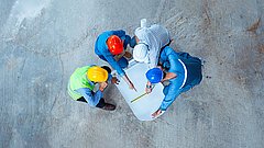 Drei Bauarbeiter mit Schutzhelmen und Warnwesten betrachten einen Bauplan auf einem Betonboden