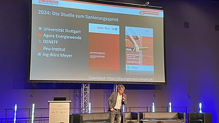 Mann mit Mikrofon auf Bühne vor Leinwand mit Präsentation zu Sanierungssprint 2024 und Logos mehrerer Institute