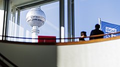 Das Bild zeigt den Berliner Fernsehturm im Hintergrund, gesehen durch ein Fenster aus einem Gebäude im Vordergrund (mit unscharfen Personen und einer blauen dena-Fahne rechts). 