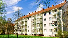 Mehrfamilienhaus mit fünf Etagen, weißen Wänden und Balkonen, umgeben von Bäumen und Rasenfläche. Blauer Himmel mit Wolken.