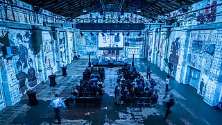 Die Eventlocation von innen, blau erleuchtet und mit dem Graphic recording an die Wand projiziert ist zu sehen.