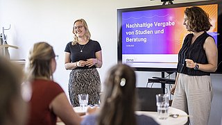 Zwei Frauen stehen vor einem Bildschirm mit der Überschrift 'Nachhaltige Vergabe von Studien und Beratung', mehrere Personen sitzen an einem Tisch und hören zu.