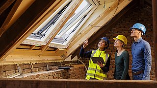 Drei Personen mit Schutzhelmen begutachten den Dachausbau eines Gebäudes, eine Fachkraft erklärt etwas und zeigt nach oben.