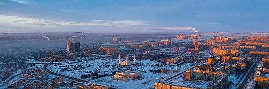 Luftaufnahme einer Stadt im Winter mit schneebedeckten Gebäuden, links renovierte und rechts unrenovierte Wohnblöcke, im Hintergrund Industrieanlagen und rauchende Schornsteine.
