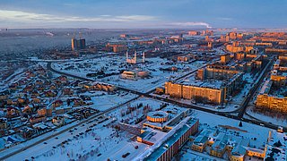 Luftaufnahme einer Stadt im Winter mit schneebedeckten Gebäuden, links renovierte und rechts unrenovierte Wohnblöcke, im Hintergrund Industrieanlagen und rauchende Schornsteine.