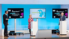 Drei Personen auf einer Bühne, zwei stehen links und rechts, eine in der Mitte. Links ein Mann in schwarzem Pullover, rechts ein Mann in Anzug. In der Mitte eine Frau in rotem Kleid mit weißen Punkten. Zwei Bildschirme zeigen 'enerthon' und 'INNOVATIONSPROJEKTE'.