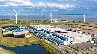 Luftaufnahme eines großen Rechenzentrums mit mehreren Gebäuden und Kühlsystemen, umgeben von Windkraftanlagen und einem Wasserlauf.