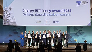 Gruppe von Personen auf der Bühne bei der Verleihung des Energy Efficiency Award 2023. Im Hintergrund ein Banner mit dem Text 'Energy Efficiency Award 2023 Schön, dass Sie dabei waren!' und Logos von dena und KFW.