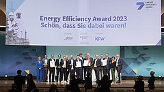 Gruppe von Personen auf der Bühne bei der Verleihung des Energy Efficiency Award 2023. Im Hintergrund ein Banner mit dem Text 'Energy Efficiency Award 2023 Schön, dass Sie dabei waren!' und Logos von dena und KFW.