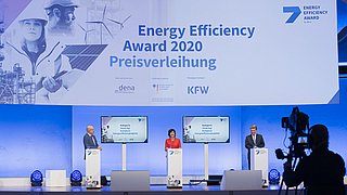 Drei Personen stehen an Pulten während der Preisverleihung des "Energy Efficiency Award 2020". Auf Bildschirmen sind Kategorien angezeigt. Im Hintergrund sind Grafiken von Windkraftanlagen und Solarpanelen.
