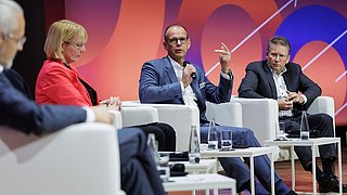 Eine Gruppe von Fachleuten sitzt auf einer Bühne in weißen Sesseln und diskutiert lebhaft. Ein Mann mit Mikrofon spricht, während die anderen aufmerksam zuhören. Der Hintergrund zeigt ein modernes, farbiges Design mit Pfeilen.