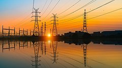Strommasten und Hochspannungsleitungen vor einem Sonnenuntergang, mit Spiegelung im Wasser.