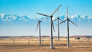 Windräder auf einer trockenen, flachen Landschaft vor schneebedeckten Bergen unter blauem Himmel.