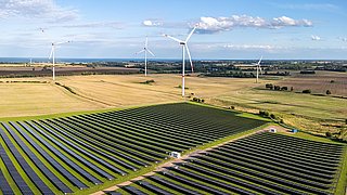Luftaufnahme eines großen Solarparks mit mehreren Reihen von Solarmodulen auf einem Feld, umgeben von Windrädern und landwirtschaftlichen Flächen unter blauem Himmel.