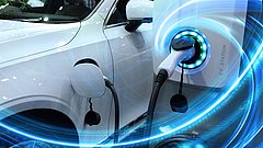 Ein weißes Elektroauto an einer Ladestation mit einem Ladekabel, das in die Fahrzeugöffnung eingesteckt ist. Die Ladestation zeigt 'EV STATION'.