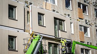 Ein schwebendes Fassadenteil wird von Bauarbeitenden in die richtige Position gebracht.