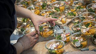 Eine Hand platziert eine Glas-Schüssel mit buntem Gemüse, darunter Brokkoli und Radieschen, auf einem Tisch voller ähnlicher Schalen.