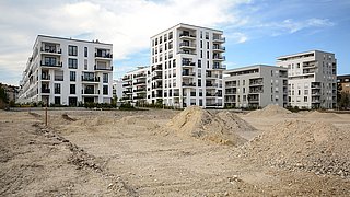 Moderne Wohngebäude mit mehreren Etagen, Balkonen und großen Fenstern, umgeben von unbebautem Gelände mit Erdhügeln.
