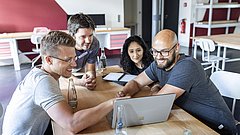 Vier Personen sitzen an einem Holztisch und schauen auf einen Laptop, der auf dem Tisch steht, in einem modernen Büro mit roten und weißen Wänden.