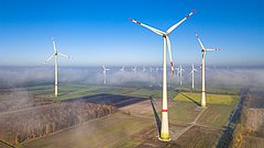 Luftaufnahme von Windkraftanlagen in einer ländlichen Umgebung, mehrere große Windturbinen mit roten Markierungen auf den Rotorblättern, umgeben von Feldern und Nebel.